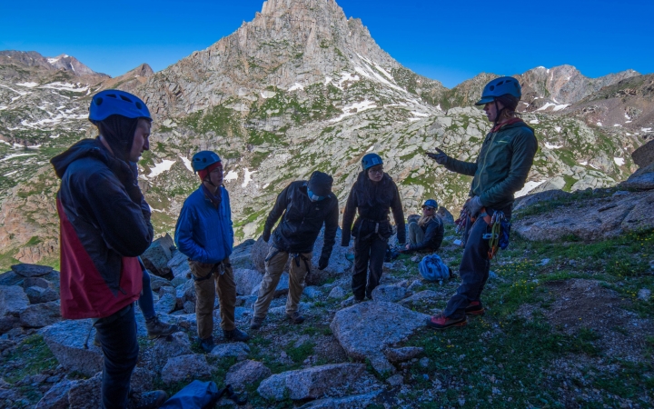 A group of people listen to an instructor speak. They are all wearing helmets, and there is a rocky mountain behind them.